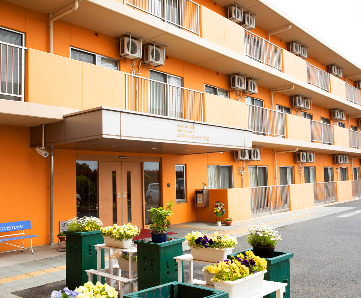 オレンジ色の外壁が特徴的な介護施設の玄関・車寄せ。花壇が配置された明るい出入口